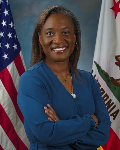 U.S. Senator Laphonza Butler, wearing a blue dress with arms crossed, standing against blue backdrop next to U.S. and California flags