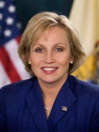 Kim Guadagno smiles in blue suit in front of American flag