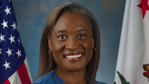 U.S. Senator Laphonza Butler, wearing a blue dress with arms crossed, standing against blue backdrop next to U.S. and California flags