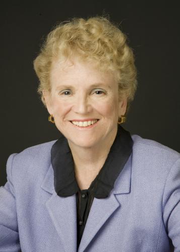 Susan Rose smiles in front of black background wearing blue gray suit jacket and a black button down with a rounded collar